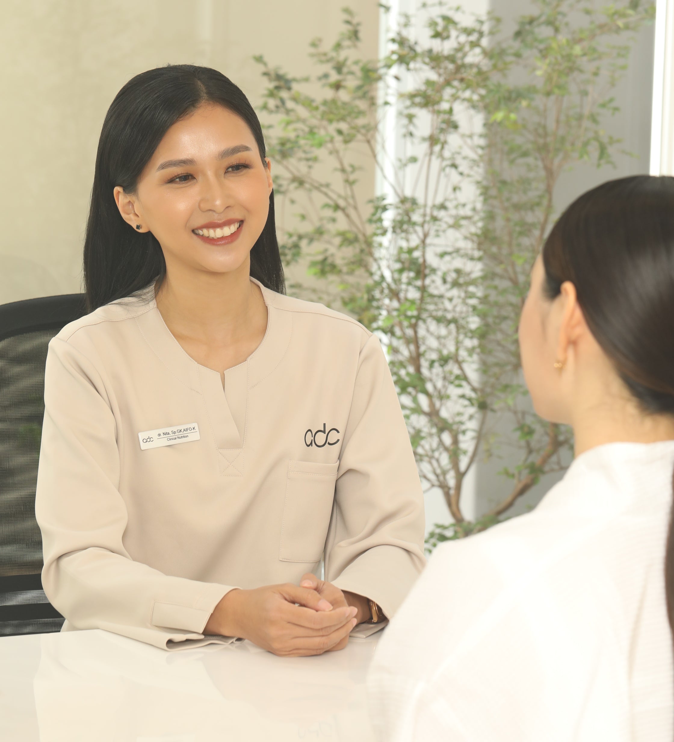 Woman in a beige sweater with a logo standing next to another person in a white coat, possibly in a medical or clinical setting.