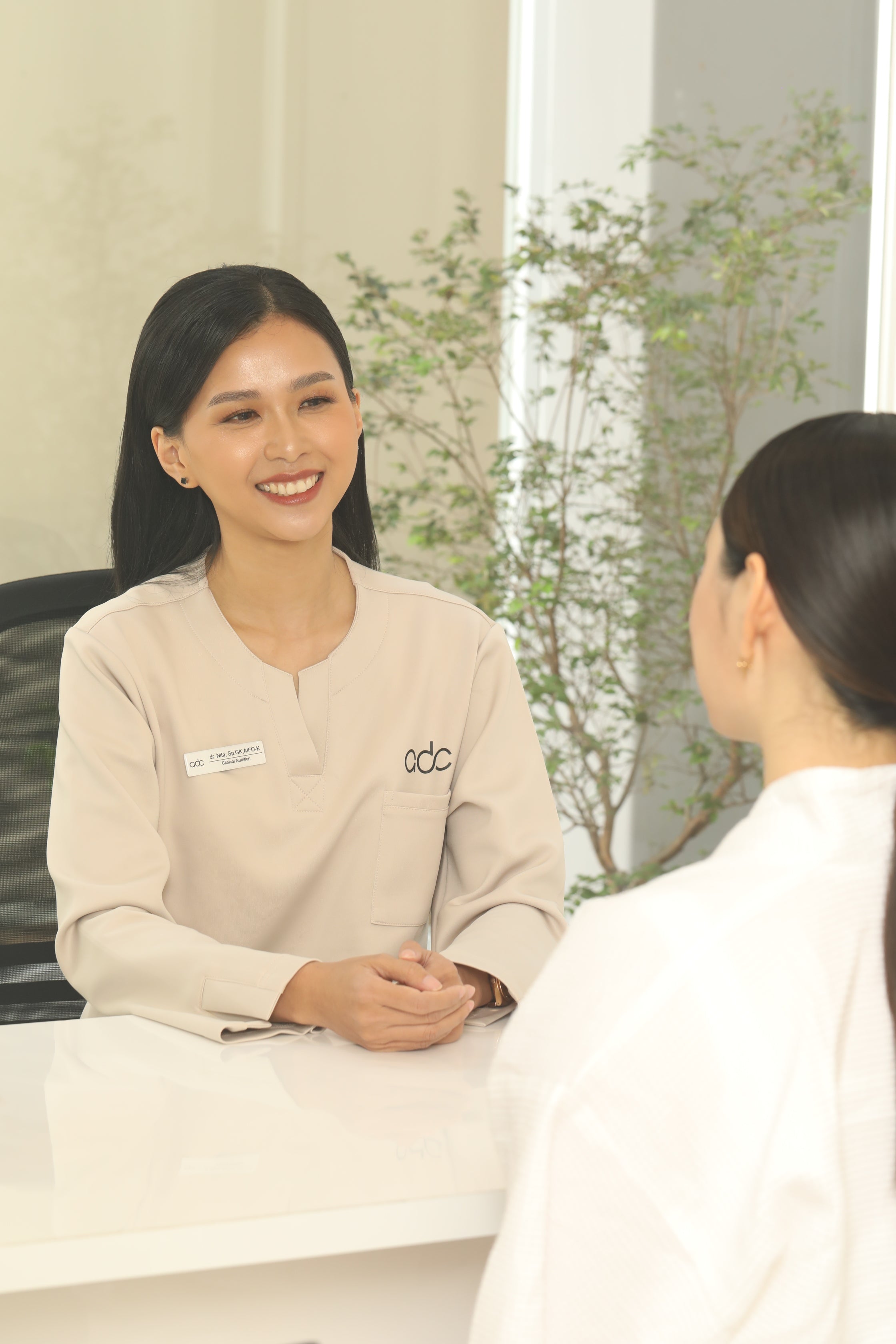 Woman in a beige sweater with a logo standing next to another person in a white coat, possibly in a medical or clinical setting.